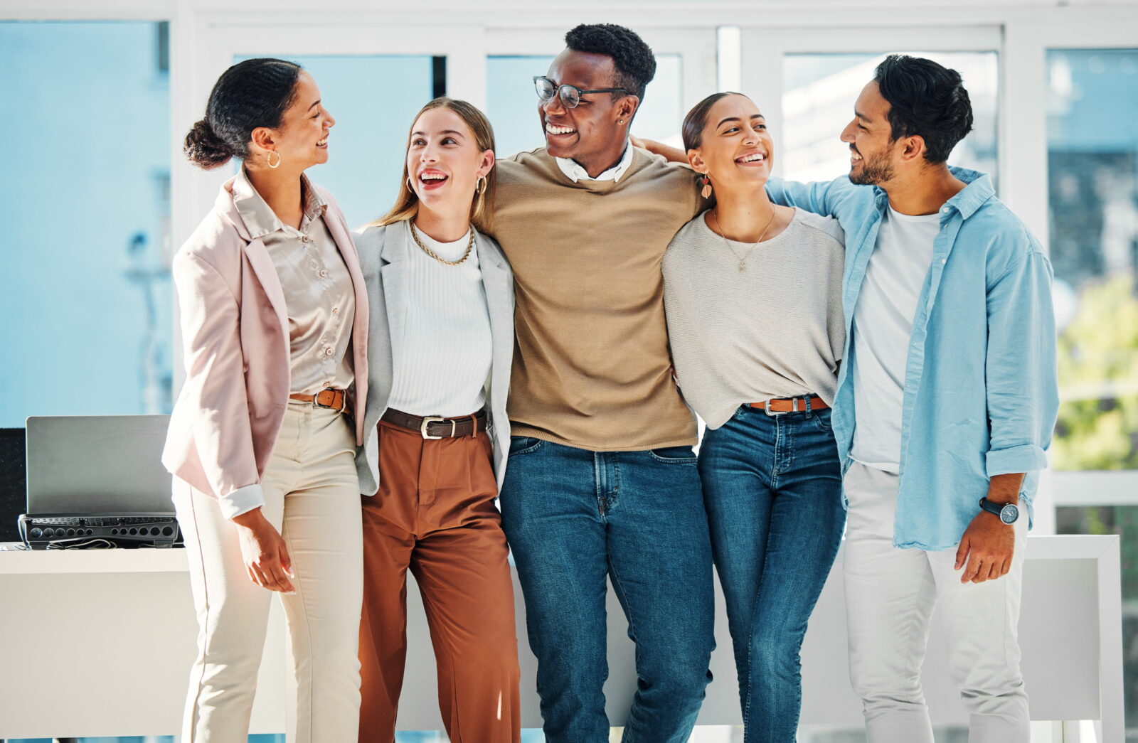 Five diverse coworkers stand together with arms around each other, smiling in a bright, modern office.