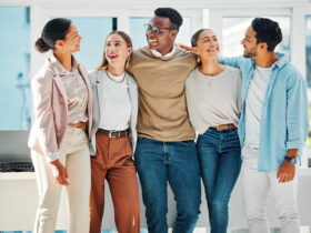 Five diverse coworkers stand together with arms around each other, smiling in a bright, modern office.