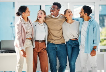 Five diverse coworkers stand together with arms around each other, smiling in a bright, modern office.