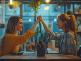 Two women in a cozy cafe share a high-five and smile at each other over a table with laptops and art supplies nearby.