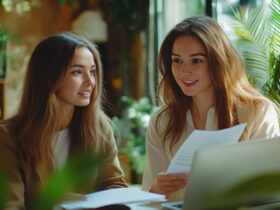 Two young women sit at a sunlit café table, looking over papers and a laptop together.