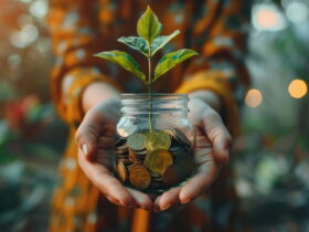 Hands holding a glass jar filled with coins, with a small plant sprouting from the top.