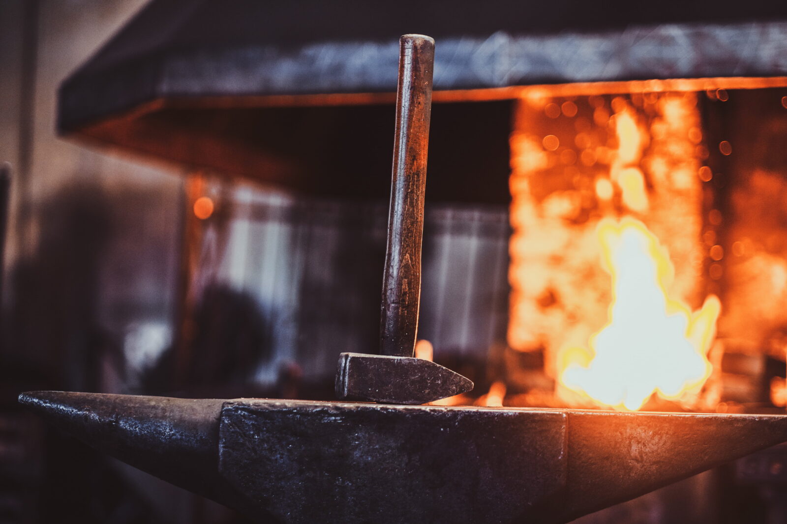 Metal rod resting on an anvil with bright orange forge flames in the background, blacksmithing scene