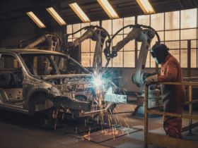 Industrial welding robot arm sparks as a worker in orange overalls watches near a car frame.