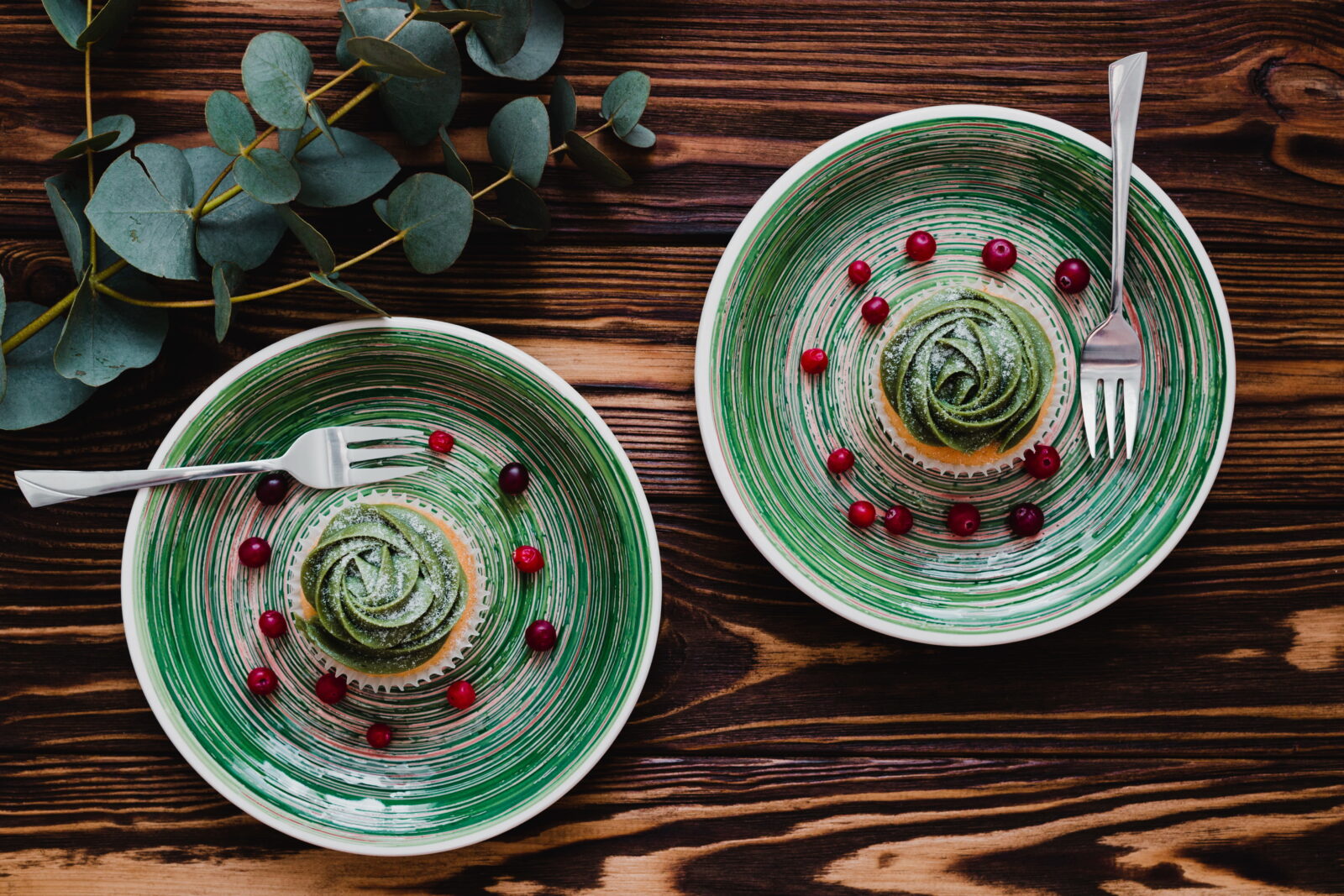 Top-down view of green-striped plates holding a matcha swirl dessert with scattered red berries on a dark wooden table, with cutlery nearby and eucalyptus on the side.”,