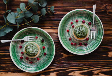 Top-down view of green-striped plates holding a matcha swirl dessert with scattered red berries on a dark wooden table, with cutlery nearby and eucalyptus on the side.”,