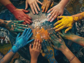 Multiple hands covered in bright paint—blue, red, yellow, and orange—reaching toward and resting on a painted blue globe, symbolizing teamwork and creativity on a collaborative art project.