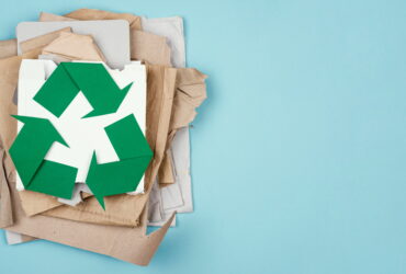 Pile of paper scraps and cardboard with a large green recycling symbol on top against a blue background, conveying recycling theme.