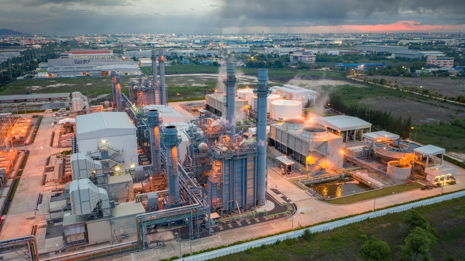 A large industrial plant with tall chimneys and piping, steam rising, set against a distant cityscape at dusk.