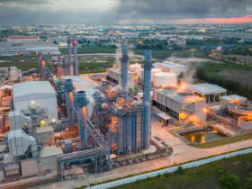 A large industrial plant with tall chimneys and piping, steam rising, set against a distant cityscape at dusk.