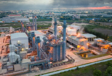 A large industrial plant with tall chimneys and piping, steam rising, set against a distant cityscape at dusk.