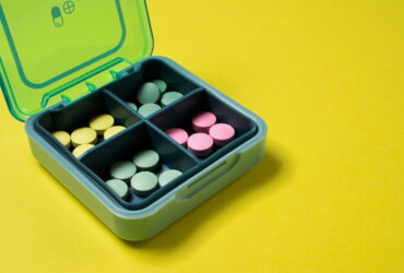 Open turquoise pill organizer with pastel yellow, green, and pink pills in its compartments on a bright yellow background moment