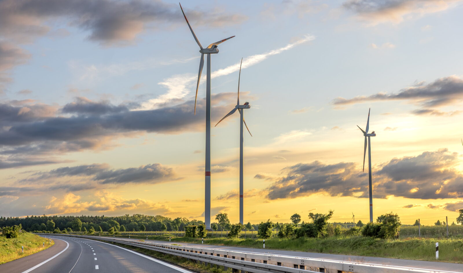 Wind turbines towering over a rural road at sunset, with a grassy field and trees in the distance.