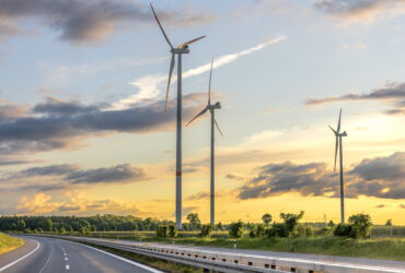 Wind turbines towering over a rural road at sunset, with a grassy field and trees in the distance.