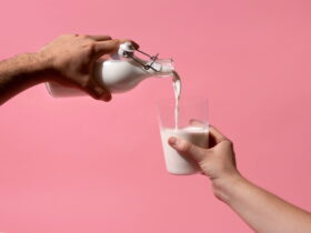 Two hands pour milk from a glass bottle into a glass against a pink background.