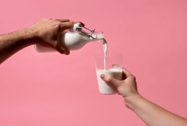 Two hands pour milk from a glass bottle into a glass against a pink background.