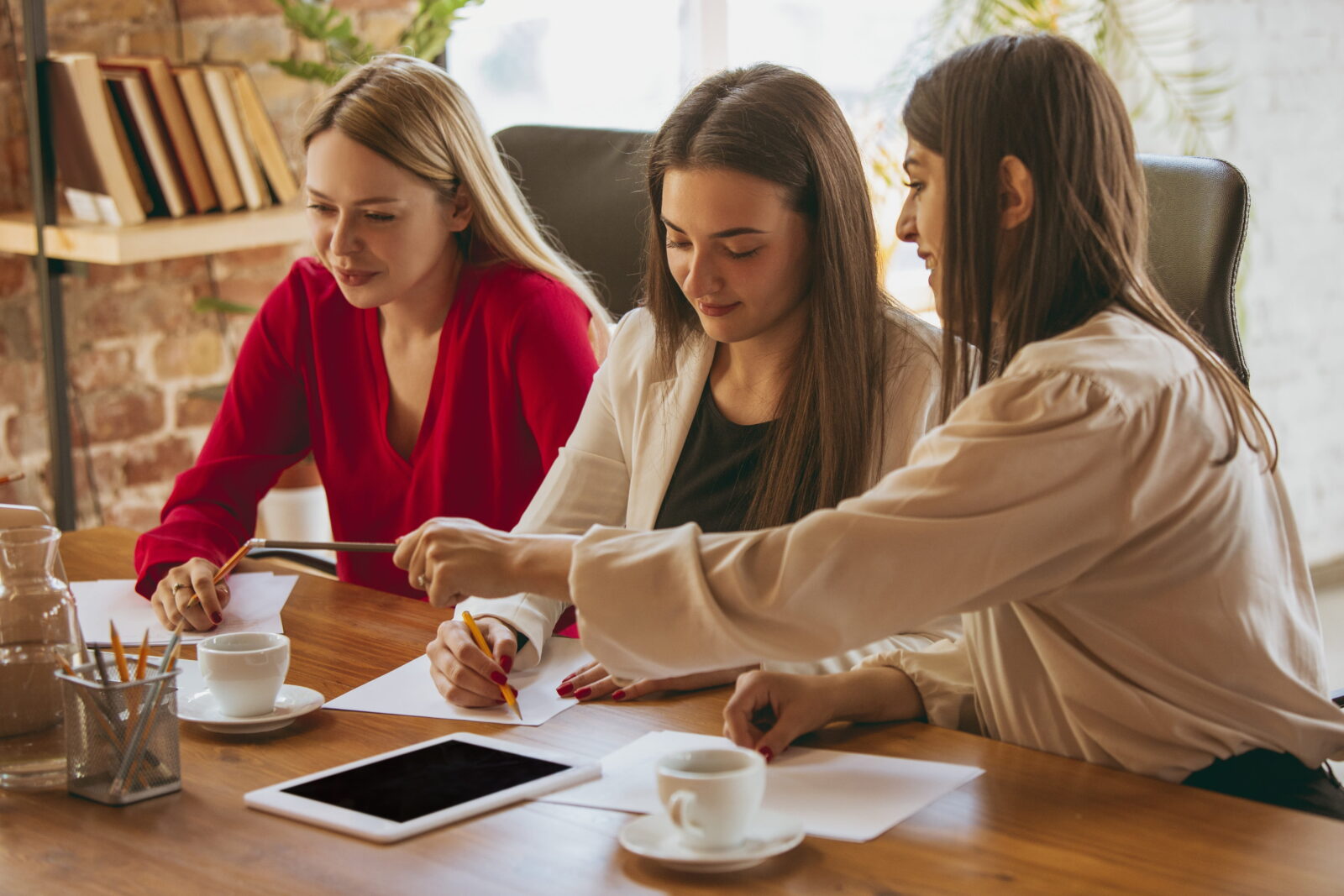 Three women sit at a wooden table sketching on paper, with pencils, near a coffee cup and tablet in a bright, casual meeting space.