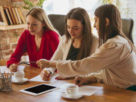 Three women sit at a wooden table sketching on paper, with pencils, near a coffee cup and tablet in a bright, casual meeting space.