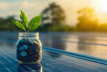 Jar filled with coins with a green sprout growing from the top, set on a reflective surface with solar panels in the background.